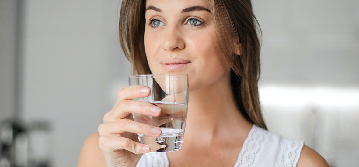 White woman wearing white clothes and holding a glass of water to hydrate herself for hair growth.