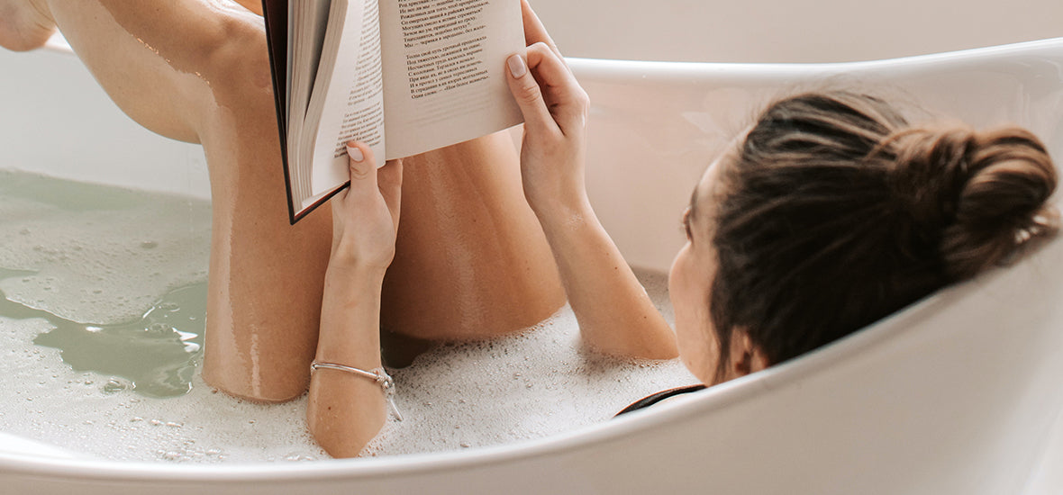 A woman lying in a white bathtub, surrounded by bubbles, with her legs in the air while reading a book for her evening routine for sleep.