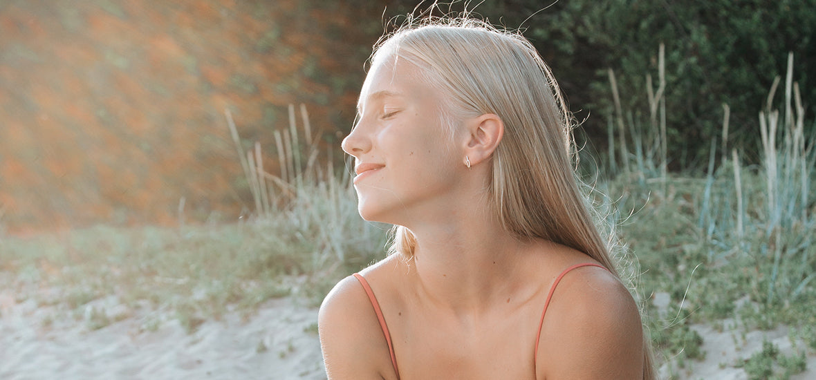 Pale woman with long white-blonde hair, sitting on a beach, holding a drink, with her eyes closed and smiling at the sunlight.