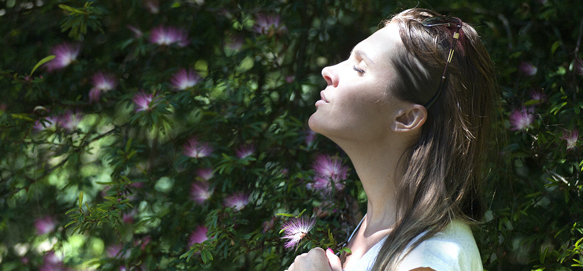 Woman standing with her eyes closed, facing the sun, with purple flowers in the background, lowering her hormonal stress.