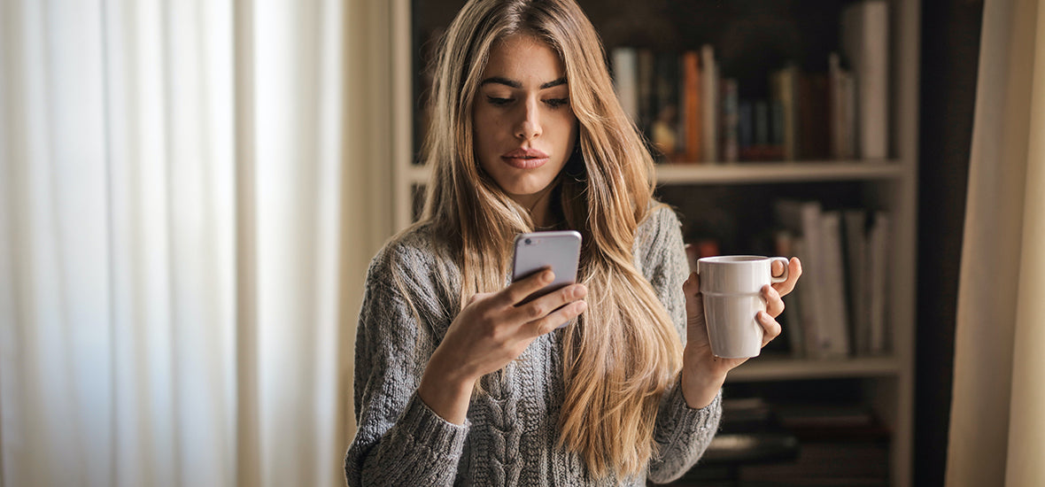 White woman with long, blonde hair in a grey jumper, holding a cup of coffee and on her phone because of bedtime procrastination.