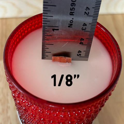 A red soy candle by The Glow Co. with a trimmed wooden wick. A ruler is measuring its wick length at 1/8 inch. 