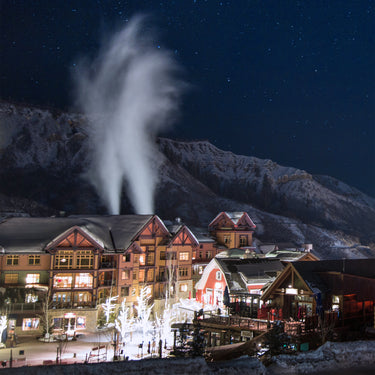 A snow covered lodge is illuminated by lights at night with a dark starry sky and mountain backdrop.