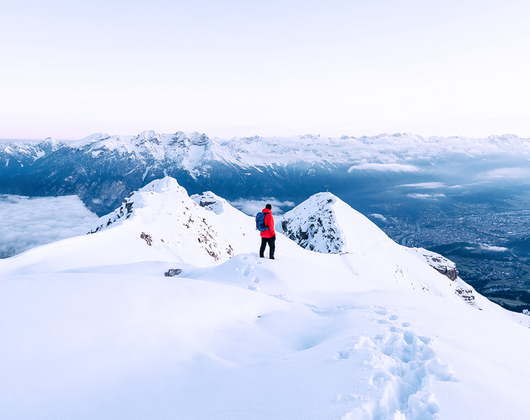 Tracks in the snow leading to a person on a mountain top with a red jacket contrasting to an epic blue horizon with a mountain range in the distance.