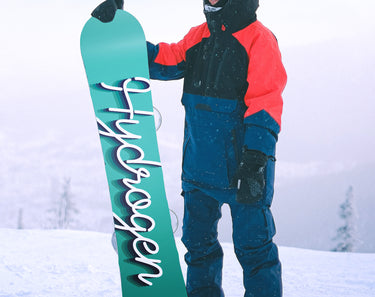 A snowboarder standing on a mountain top in choppy snow, shows off the back of his snowboard which reads Hydrogen in a cursive script.