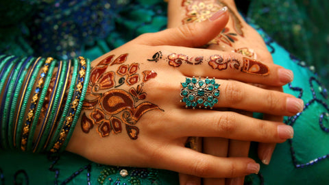 Henna hands is a transnational arabic accessory painted onto hand for many occasions, especially weddings. pictures is a woman's hands with aqua bracelets and ring folded over an aqua dress over her knee.