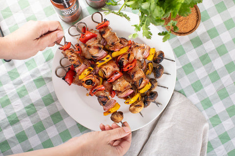A plate of cooked super skewers looking juicy and delicious, with 2 hands picking up one skewer off the plate