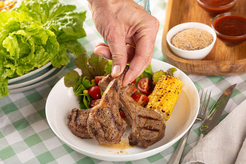 A hand picking up a barbecued lamb cutlet from a plate of juicy lamb cutlets, BBQ corn and garden salad.