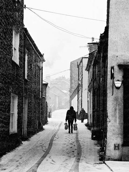 Black and white image showing a person walking uphill on a snowy road in Kendal