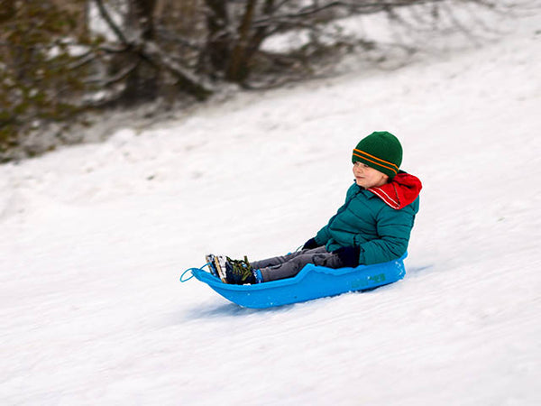 Image shows a snow-covered slope with a child going downhill on a sledge