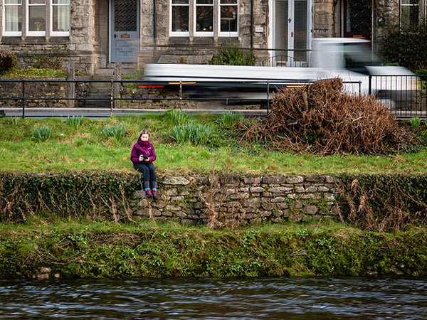 Image shows a girl with a camera sitting on a wall overlooking the river, as traffic passes behind