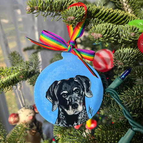 Acrylic painting of a black and grey dog on a blue background, painted on a Christmas ornament. The ribbon is rainbow. 