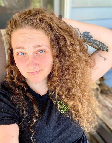 Selfie of Amanda in the summertime. Her curly dirty blonde hair is down around her shoulders, her left arm is up behind her head. There is a tattoo visible on her arm and a twinkle in her eye.