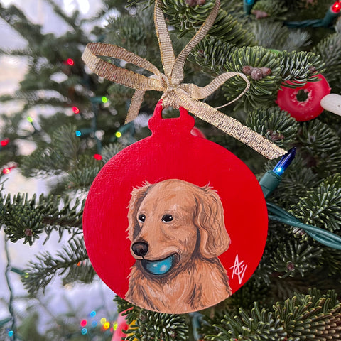 Acrylic painting of a golden retriever with a blue ball in his mouth against a red background. The painting is on a Christmas ornament.