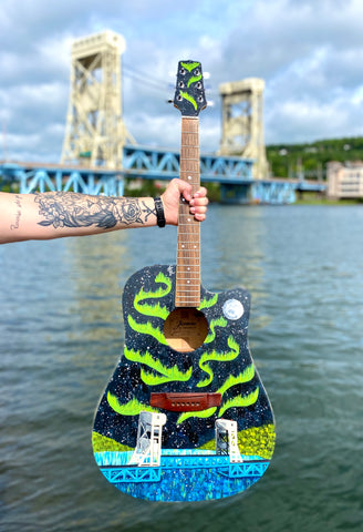 Hand painted guitar depicting the Portage Lake Lift Bridge with Northern Lights behind it. The guitar is being held in front of the real Portage Lake Lift Bridge in Houghton Michigan.