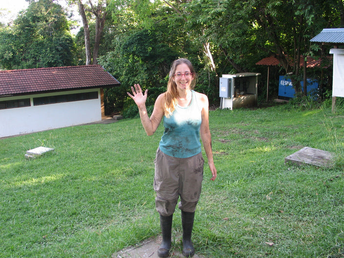 Kris is shown waving at the camera with long, red hair in a low ponytail wearing a light turquoise tank top, gray pants, and black mud boots and spotted with mud is standing in a grassy yard surrounded by trees. 