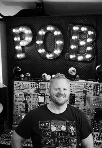 Patrick standing in front of a large modular synthesizer system. On top of the system is a large illuminated sign that says "POB". A black cat sits on top of the sign. 