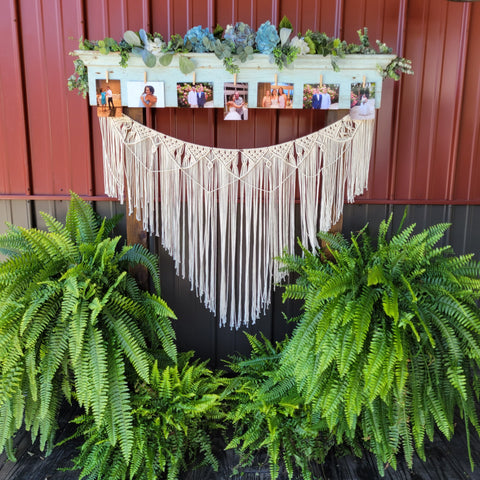 Gorgeous antiqued aqua mantel with a full greenery garland across the top and photographs all along the front. A large beautiful macramé the width of the mantel hanging below. Surrounded by ferns.