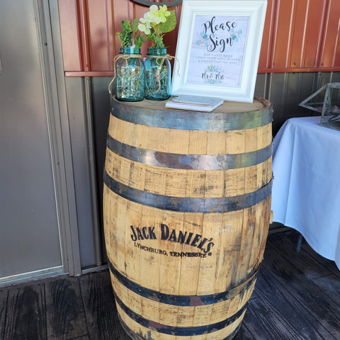 Jack Daniels whiskey barrel with framed sign "Please Sign our Guest Book." Flower arrangement in blue Ball mason jars.