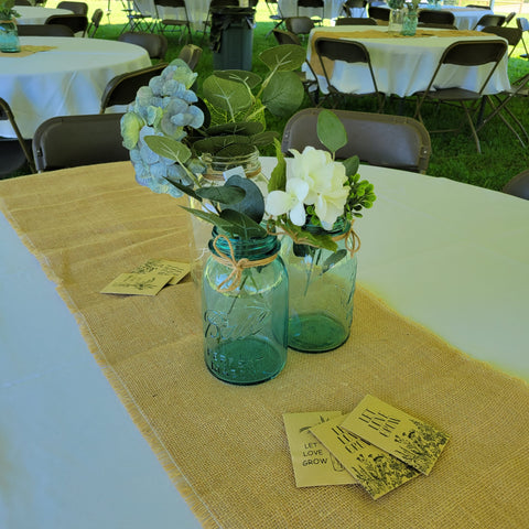 Guest table centerpieces: kraft paper packets of wildflower seeds, blue Ball mason jars with green and white flowers, burlap runner over white tablecloth.