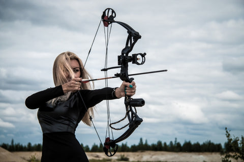 Young blonde female wields a compound bow and looks into bow sight ready to shoot.
