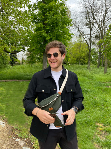 Homme vingtaine portant un sac banane et un étui à lunette vert foncé dans un parc