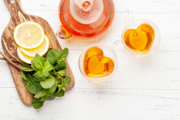 Steeped tea in two heart-shaped clear mugs next to a cutting board with sliced lemon and fresh mint leaves.