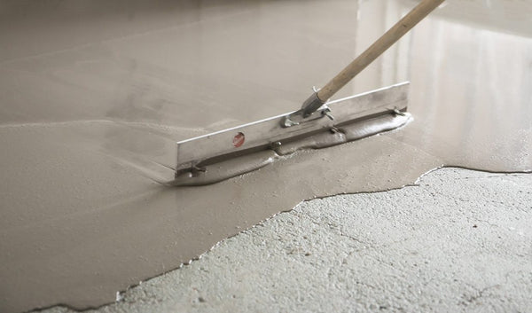 A gauge rake pushing a self-leveling concrete material across a floor surface.