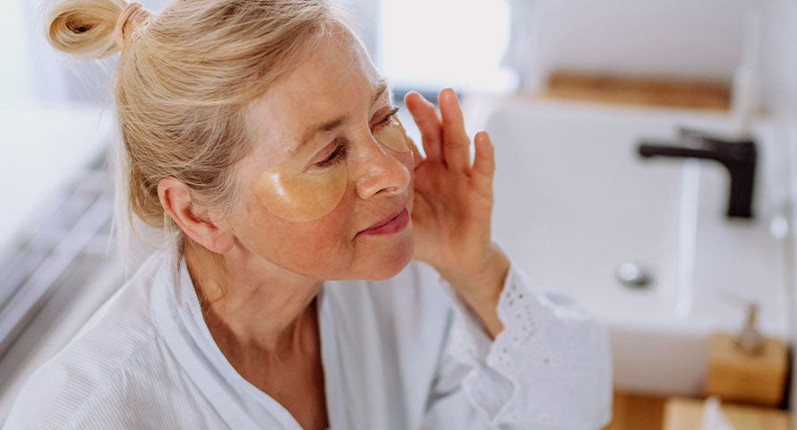 Alt Description: A woman delicately applying a hydrating eye gel patch under her eye, depicting a focused and gentle step in her skincare regimen.