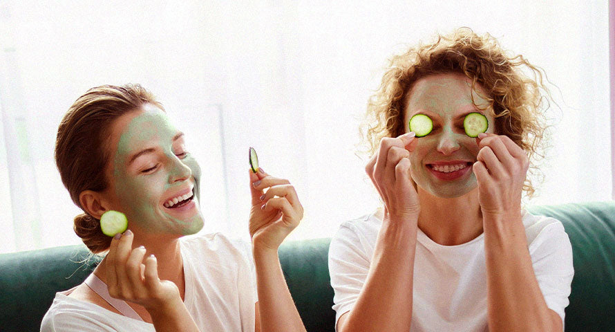 Image of a woman with a facial mask applied, holding up two slices of cucumber in front of her eyes, embodying a relaxed and rejuvenating skincare routine.