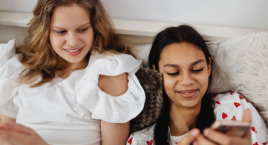 Two friends of diverse ethnic backgrounds sitting closely on a sofa, sharing a joyful moment as they look at a phone together and smile.