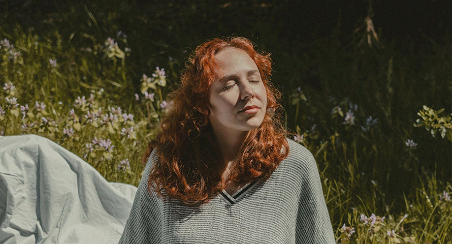 A woman standing outside, closing her eyes with a serene expression, as she tilts her face towards the sunlight, enjoying the warmth and light on her skin.