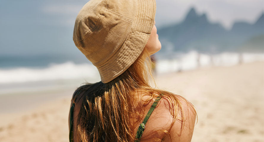 Rear view of a woman wearing a sun hat at the beach, looking out over the ocean, embodying peaceful solitude.