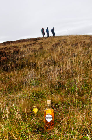 Bottle of river rock whisky placed on the grass with walkers on a hill in the background