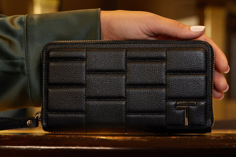A hand holding the Trevony Zipped Wallet in black lambskin with a distinctive woven pattern and a silver T-shaped clasp, showcasing the wallet's elegant design and craftsmanship.
