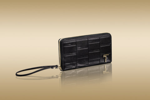 A Trevony Zipped Wallet in textured lambskin with a wrist strap and a prominent gold T-clasp, set against a warm, reflective golden background.