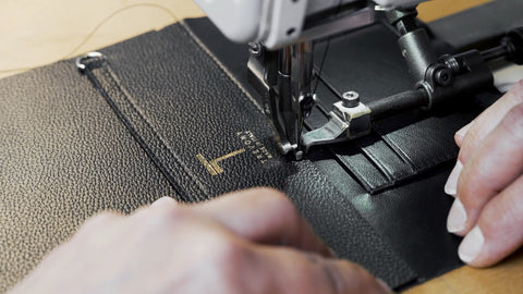 Close-up view of a sewing machine stitching a black Minerva handbag with the golden Trevony logo, demonstrating the brand's precision in manufacturing luxury goods.