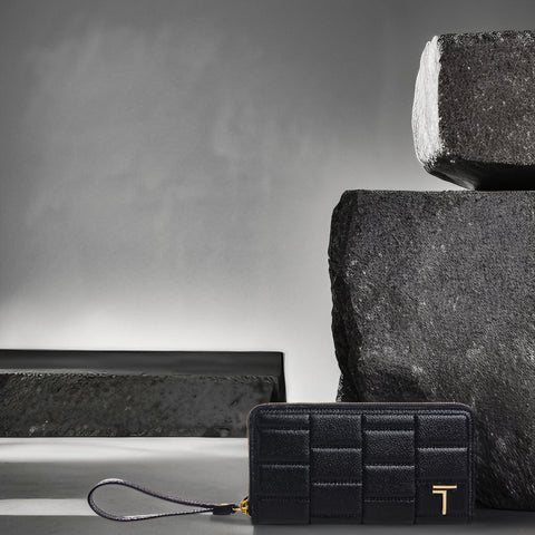 A sophisticated black Trevony women's zipped wallet, featuring a textured surface with horizontal ridges and a prominent gold-tone Trevony logo. The wallet has a wristlet strap for easy carrying and is displayed against a backdrop of artistic, angular stone structures, conveying a sense of luxury and modern design.