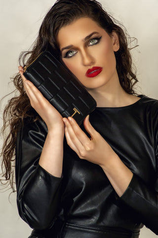 A woman with dramatic makeup and red lipstick holding a Trevony Zipped Wallet in black lambskin against her black leather dress, with the wallet's woven pattern and gold T-clasp on display.