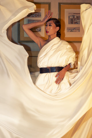 A model showcases the Trevony Truth Oversized Belt, cinching a flowing white gown at the waist. The belt's prominent buckle and textured surface provide a bold contrast to the soft fabric of the dress. She poses dramatically with one hand raised to her forehead, embodying confidence and elegance, against a backdrop of warm-toned wall art.