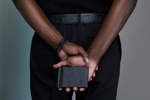 A sophisticated individual in a tailored black outfit holds the Trevony Vinny Bill Clip Wallet. The wallet's premium black lambskin leather texture is visible, accented with a subtle embossed 'T' logo, signifying the Trevony brand. The wallet conveys understated luxury, complemented by the wearer's stylish beaded bracelet.
