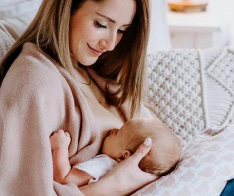 Nouvelle maman qui sourit en allaitant son jeune bébé, tous deux confortablement installés sur un coussin d'allaitement Neka en écalles de sarrasin.