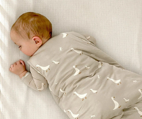 Bébé qui dort confortablement dans son sac de nuit, aussi appelé gigoteuse, à motif d'oie de la marque Gunamuna