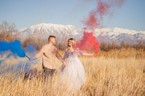 Man holding blue smoke bomb and pregnant woman holding pink smoke bomb for gender reveal