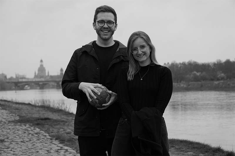 Tom und Linda stehen in Dresden am Elbufer. In der Hand hält Tom einen Handball und im Hintergrund sieht man die Frauenkirche.