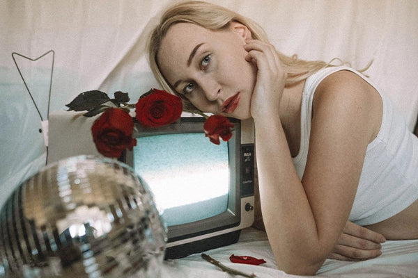 Girl with blonde hair leaning against small TV with roses on it next to disco ball