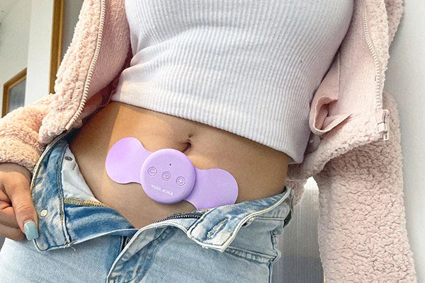 Person with white top and pink jacket wearing purple Aura pain relief device on stomach with blue jeans undone to show product