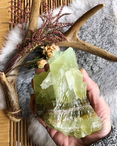 Large green calcite chunk held in hand with wicker, fur and deer antler blurred in the background.