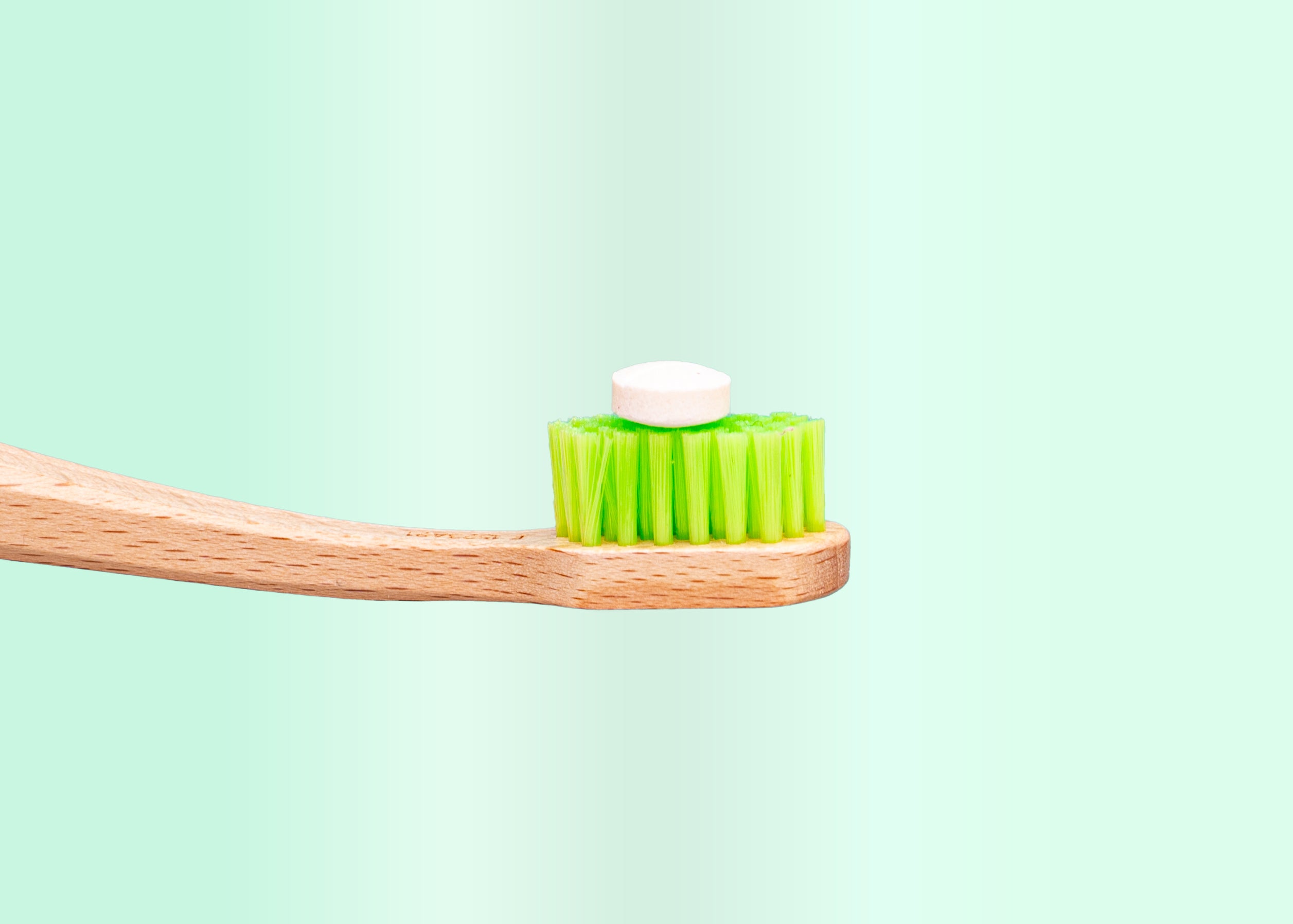 A wooden toothbrush with green bristles and a white tablet on top.