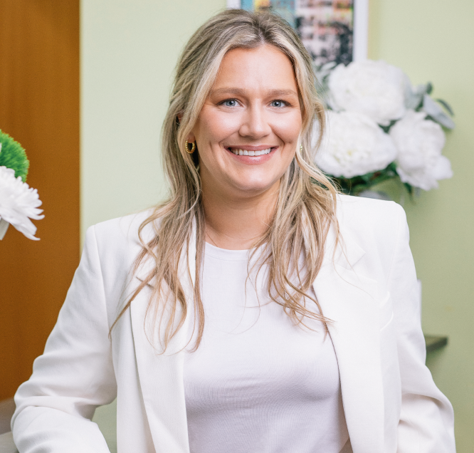 Woman smiling in a white blazer with flowers in the background.